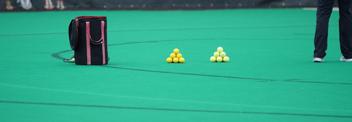 A sports field features a black and pink sports bag, two pyramids of yellow balls, and a portion of a person in dark athletic gear. The background displays a banner with the Under Armour logo.