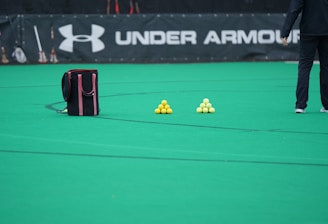 A sports field features a black and pink sports bag, two pyramids of yellow balls, and a portion of a person in dark athletic gear. The background displays a banner with the Under Armour logo.