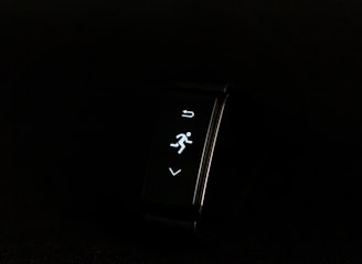A digital fitness tracker with a black band is displayed against a dark background. The screen features minimalistic white icons for navigation and an exercise mode, including an arrow, a running figure, and a downward-pointing arrow.