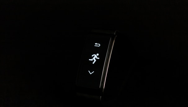 A digital fitness tracker with a black band is displayed against a dark background. The screen features minimalistic white icons for navigation and an exercise mode, including an arrow, a running figure, and a downward-pointing arrow.