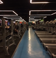 A spacious gym with multiple rows of treadmills and weightlifting benches on either side. The lighting features geometric patterns on the ceiling, creating a modern and organized ambiance. People are actively using the equipment, engaging in fitness activities.
