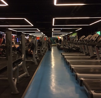 A spacious gym with multiple rows of treadmills and weightlifting benches on either side. The lighting features geometric patterns on the ceiling, creating a modern and organized ambiance. People are actively using the equipment, engaging in fitness activities.