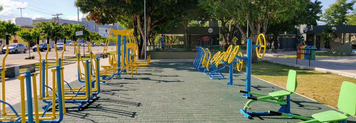 An outdoor fitness area featuring a variety of exercise equipment, including stationary bikes and other workout stations. The equipment is primarily blue and yellow, arranged neatly on a paved surface. Surrounding the area are trees providing shade, with a few parked cars and buildings visible in the background.