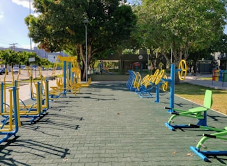 An outdoor fitness area featuring a variety of exercise equipment, including stationary bikes and other workout stations. The equipment is primarily blue and yellow, arranged neatly on a paved surface. Surrounding the area are trees providing shade, with a few parked cars and buildings visible in the background.