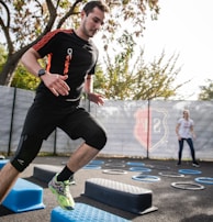A man is actively participating in an outdoor fitness session, jumping over blue platforms with agility. Two other people in the background are also engaging in exercise, with rings scattered on the ground. The area is fenced and surrounded by trees, suggesting a park or recreational setting.