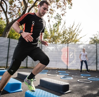 A man is actively participating in an outdoor fitness session, jumping over blue platforms with agility. Two other people in the background are also engaging in exercise, with rings scattered on the ground. The area is fenced and surrounded by trees, suggesting a park or recreational setting.