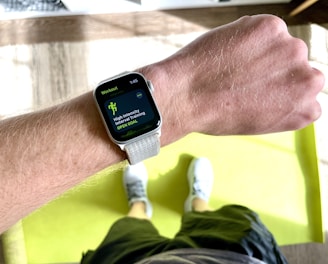 A close-up view of a person's wrist wearing a smartwatch displaying a workout mode for High Intensity Interval Training. The background includes a bright green exercise mat on a wooden floor. The person is wearing white athletic shoes and dark shorts.