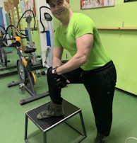 A man wearing a bright green athletic shirt, black pants, and a cap is standing in a gym. He has one foot on a small platform and is smiling at the camera. In the background, there are exercise bikes and artwork on the green walls.