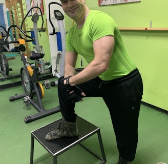A man wearing a bright green athletic shirt, black pants, and a cap is standing in a gym. He has one foot on a small platform and is smiling at the camera. In the background, there are exercise bikes and artwork on the green walls.