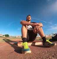 A person wearing a black athletic outfit is seated on a wooden platform in an outdoor sports field. The individual is relaxed, with arms resting on knees. The field is expansive with a clear blue sky and scattered clouds above. A brightly colored running shoe and athletic gear are visible, along with a water bottle and a blue backpack placed beside the person. Lampposts and residential buildings can be seen in the background.