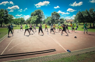 A group of people are exercising outdoors on a concrete court, surrounded by green spaces and trees. They are standing in a formation, appearing to engage in an organized fitness class. Each participant wears athletic clothing, and several carry exercise equipment such as dumbbells and ropes. The sky is bright blue with scattered fluffy clouds, creating a vibrant and energetic atmosphere.