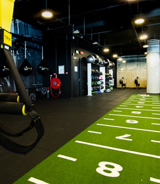 A modern gym space featuring a green track laid out on the floor with numbered lanes. TRX suspension trainers hang prominently in the foreground. The gym is equipped with various exercise equipment, including kettlebells and punching bags. In the background, there is a FIT sign near a reception desk under a low-ceilinged industrial design.