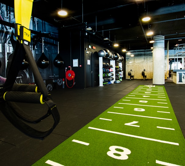 A modern gym space featuring a green track laid out on the floor with numbered lanes. TRX suspension trainers hang prominently in the foreground. The gym is equipped with various exercise equipment, including kettlebells and punching bags. In the background, there is a FIT sign near a reception desk under a low-ceilinged industrial design.