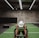 A person in athletic wear is pushing a sled on green turf inside a dimly lit gym with 'Global Fitness' written on the wall. The setting includes exercise equipment and industrial elements, such as visible metal beams and a rolled-up garage door.