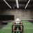 A person in athletic wear is pushing a sled on green turf inside a dimly lit gym with 'Global Fitness' written on the wall. The setting includes exercise equipment and industrial elements, such as visible metal beams and a rolled-up garage door.