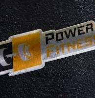 A metal plaque with the text 'POWER FITNESS' is set against a textured black background. The design includes a stylized dumbbell icon adjacent to the text, with a modern and sleek appearance.