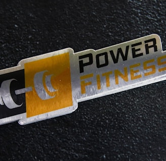 A metal plaque with the text 'POWER FITNESS' is set against a textured black background. The design includes a stylized dumbbell icon adjacent to the text, with a modern and sleek appearance.