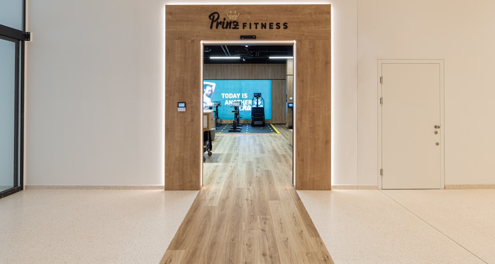 A modern fitness center entrance with wooden framing and the sign 'PRIME FITNESS' above the doorway. The interior features a spacious gym area with workout equipment visible and a motivational banner on the back wall. The flooring transitions from glossy tiles to a wood finish leading into the facility.