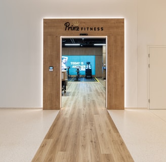A modern fitness center entrance with wooden framing and the sign 'PRIME FITNESS' above the doorway. The interior features a spacious gym area with workout equipment visible and a motivational banner on the back wall. The flooring transitions from glossy tiles to a wood finish leading into the facility.