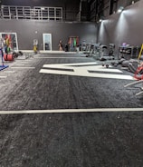 A spacious gym with a black and white color scheme featuring various fitness equipment. On the left, numerous weight plates and bars are organized on racks. In the center, a large open workout area is visible with a textured floor and white directional markings. To the right, there are treadmills, stationary bikes, and other workout machines along with large exercise balls. Exercise bands and weightlifting belts are hanging on hooks at the back wall. A single person is seen near a door, adding a human element to the scene.