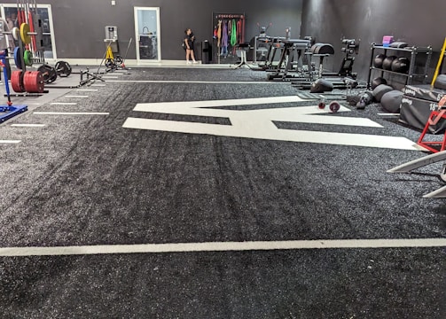 A spacious gym with a black and white color scheme featuring various fitness equipment. On the left, numerous weight plates and bars are organized on racks. In the center, a large open workout area is visible with a textured floor and white directional markings. To the right, there are treadmills, stationary bikes, and other workout machines along with large exercise balls. Exercise bands and weightlifting belts are hanging on hooks at the back wall. A single person is seen near a door, adding a human element to the scene.