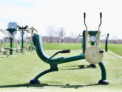 An outdoor gym with several pieces of exercise equipment arranged on a green artificial turf. The image focuses on a leg press machine in the foreground with other fitness machines visible in the background. The area is surrounded by an open, empty landscape.