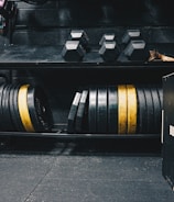 A gym setting featuring various fitness equipment, including kettlebells, stacked weight plates on a rack, dumbbells, and a medicine ball. The gym has a rugged, industrial vibe with black walls and a Bulldog Gear plyo box.