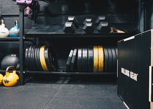 A gym setting featuring various fitness equipment, including kettlebells, stacked weight plates on a rack, dumbbells, and a medicine ball. The gym has a rugged, industrial vibe with black walls and a Bulldog Gear plyo box.