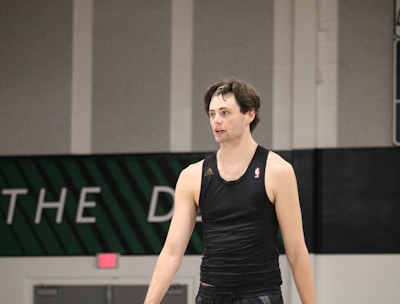 A person wearing a sleeveless black sports top is standing indoors in a gymnasium. The background includes a gray wall with green and white lettering partially visible.