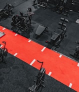 A gym setting featuring a variety of exercise equipment such as weight machines, stationary bikes, and a sled on a bright red track. The gym has a modern aesthetic with black and red color schemes.