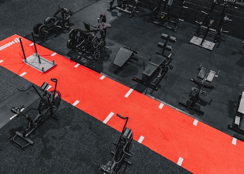 A gym setting featuring a variety of exercise equipment such as weight machines, stationary bikes, and a sled on a bright red track. The gym has a modern aesthetic with black and red color schemes.