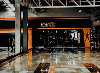 A fitness center entrance with the branding 'smart fit' illuminated above the doors. The interior has a modern design, featuring dark glass and bright orange accents. A reflective marble floor extends in front, capturing reflections from the surroundings. There are two individuals standing and walking towards the left.