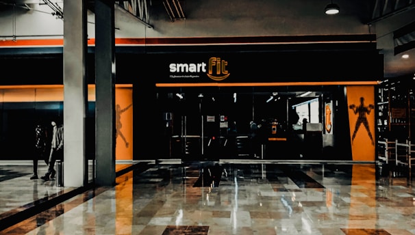 A fitness center entrance with the branding 'smart fit' illuminated above the doors. The interior has a modern design, featuring dark glass and bright orange accents. A reflective marble floor extends in front, capturing reflections from the surroundings. There are two individuals standing and walking towards the left.